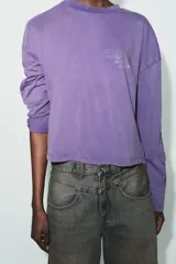 Camiseta de manga larga con corte oversize, confeccionada en algodón color violeta con efecto lavado. Presenta cuello redondo, bajo asimétrico y detalles de texto con aplicación de brillos en el pecho y las mangas.