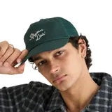 Gorro trucker verde oscuro con paneles de malla en la parte trasera, visera curva y cordón decorativo en la base de la frente. Presenta el texto "Rhythm Livin'" bordado en blanco con tipografía cursiva.