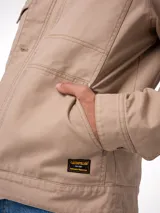 Chaqueta de canvas color beige con forro interior de franela. Presenta cierre frontal con botones metálicos, bolsillos en el pecho con solapa y bolsillos laterales. Incluye etiqueta de marca en el bolsillo inferior.