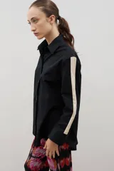 Camisa de corte holgado y largo medio, color negro con detalle de franja lateral en color crema a lo largo de las mangas. Presenta cuello clásico, cierre frontal con botones y tajos laterales en el ruedo.