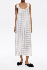Vestido largo blanco con estampado de líneas negras formando un patrón de cuadrícula. Tiene tirantes anchos con detalle de lazo en los hombros, escote redondo pronunciado en delantero y espalda, y corte línea A.