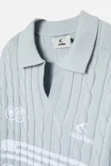 Polo de punto celeste con patrón trenzado, cuello tipo polo abierto y mangas largas. Presenta logos estampados en el pecho y diseño de estilo deportivo retro.
