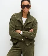 Campera de pana cotelé color verde militar, con cuello camisero, cierre frontal con botones metálicos y bolsillos frontales con solapa. Presenta diseño de sisa desplazada y cordones ajustables en el ruedo.