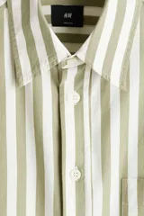 Camisa de popelina de algodón con cuello inglés, tapeta clásica, bolsillo superior abierto y canesú con pliegue en la espalda. Cuenta con mangas largas, puños ajustables con botón y bajo ligeramente redondeado. Presenta un diseño de rayas verticales en tonos verde y blanco.