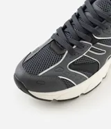Championes estilo dad sneaker de color gris oscuro, con detalles en blanco y líneas plateadas reflectivas. Combinan malla y paneles sintéticos con una suela voluminosa blanca y negra.