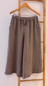 Pantalón capri color gris topo de lino con cintura elástica ajustable con cordón, bolsillos laterales y terminación con flecos.