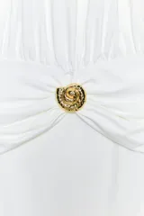 Vestido midi blanco de cuello halter, confeccionado con mezcla de poliamida. Presenta detalle de drapeado lateral con un broche metálico dorado en forma de caracola y cierre en la espalda ajustable con lazadas.