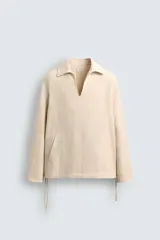 Camisa de manga larga color beige claro con textura jacquard arrugada, cuello solapa con abertura en V y bolsillo canguro frontal. Posee cordones ajustables en el bajo lateral.