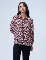 Camisa de gasa con estampado animal print en tonos marrones, violetas y naranjas. Tiene cuello camisero con ribete de puntilla negra, mangas largas abullonadas con puños elastizados y cierre frontal con botones negros.