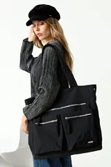 Cartera tipo shopper de gran tamaño en color negro, confeccionada en poliéster. Presenta múltiples bolsillos frontales con cierres metálicos y tiradores de cordón, además de un compartimento principal con cierre. Cuenta con correas anchas para llevar al hombro.