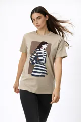 Remera de algodón color beige con cuello redondo y mangas cortas. Presenta un estampado frontal artístico de una figura femenina con detalles de studs metálicos.
