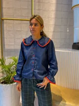 Camisa de jean azul oscuro con cuello estilo marinero y puños con volados, ambos con ribetes en color rojo. Presenta cierre frontal con botones rojos.