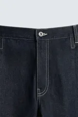 Bermuda de denim rígido color azul oscuro (sin prelavar) con corte amplio (baggy fit). Presenta pliegues frontales, trabillas ajustables en la cintura, bolsillos delanteros y bolsillos traseros de parche, todo resaltado con pespuntes a contraste.