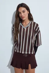 Remera de estilo deportivo con cuello en V y mangas cortas amplias. Presenta un diseño de rayas verticales en tonos marrones, blancos y líneas finas en fucsia, confeccionada en tejido de red.