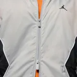 Campera rompevientos con capucha Nike Jordan, diseño color block en tonos gris claro, gris oscuro y negro. Presenta cierre frontal completo, bolsillos laterales y logo Jumpman bordado en el pecho.