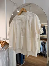 Camisa de algodón color blanco crudo, de manga corta, con diseño amplio oversize, cuello redondo con volados y cierre frontal con botones.