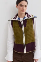 Chaleco de lana con cierre frontal completo y cuello alto. Presenta un diseño de bloques de color en tonos verde oliva, bordó y detalles en crudo y azul, con combinación de texturas en punto trenzado y punto elástico. Posee un corte asimétrico, siendo más largo en la parte posterior, y aberturas laterales en el ruedo.
