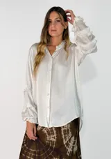 Camisa de satén color blanco hueso, con cuello mao, cierre frontal de botones y mangas largas con volados decorativos.