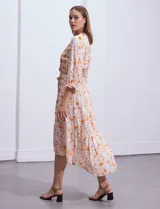 Vestido midi de gasa con estampado geométrico y floral en tonos naranja y turquesa sobre base beige. Presenta escote en V, cierre frontal con botones, mangas 3/4 con puños fruncidos y cintura elastizada con lazo de ajuste. El diseño incluye un ruedo con volado y corte asimétrico más largo en la parte posterior.