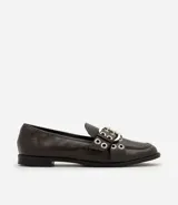 Mocasín femenino de cuero sintético color marrón oscuro, con punta redondeada y suela plana. Presenta un diseño con tiras cruzadas en el empeine decoradas con ojales metálicos y una hebilla ajustable.