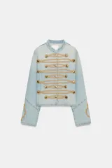 Chaqueta de mezclilla clara estilo militar, con cuello redondo, manga larga y detalles de cordones y botones dorados en el frente.