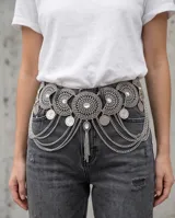 Cinto de metal plateado estilo cadena, con diseño ornamentado de inspiración étnica compuesto por placas circulares grabadas, apliques de pedrería y múltiples tiras de cadenas colgantes con dijes de monedas.