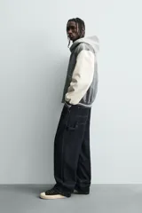 Cazadora estilo varsity o bomber, cuerpo principal gris oscuro y mangas color crema. Presenta cuello, puños y bajo en rib elástico con rayas grises y blancas. Tiene bordados en el pecho: un monograma estilizado a la izquierda y el texto "EST. 1975" a la derecha.