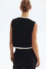 Chaleco corto negro en tejido bouclé, con cuello redondo, cierre frontal de botones dorados y bolsillos frontales decorativos ribeteados en blanco.