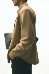 Camisa de manga larga color camel, confeccionada en sarga suave, con cuello americano y tapeta clásica con botones. Presenta canesú en la espalda y bajo redondeado.