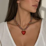 Collar corto compuesto por cuentas acanaladas en forma de corazón en tonos grisáceos y rojizos con pequeñas cuentas doradas intermedias, del cual cuelga un gran dije central en forma de corazón rojo translúcido con detalles dorados.