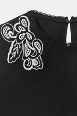 Blusa corta negra con mangas abullonadas y bordados florales en contraste de color blanco. Presenta cuello redondo con puntilla, mangas ajustables con cordones y borlas, y cierre en la espalda con abertura y botón oculto.