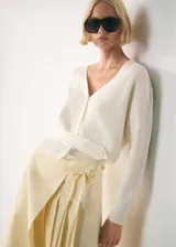 Cardigan de punto acanalado color blanco, con cuello en pico, manga larga y cierre frontal de botones. Presenta un diseño recto con una tira decorativa ajustable en la cintura con botones.
