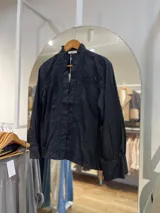 Camisa negra con cuello alto, mangas largas abullonadas y cierre frontal con botones. Presenta detalles de encaje y lentejuelas en el frente.