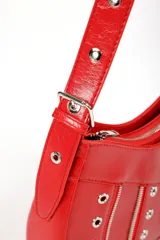 Cartera tipo baguette de color rojo con acabado brillante, decorada con múltiples cierres metálicos, ojales y dos pequeñas hebillas decorativas en el frente. Incluye un bolsillo pequeño con solapa y cierre de botón a presión, además de una correa larga ajustable.
