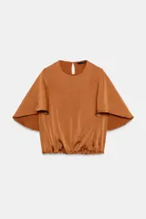 Camisa fluida color tostado de manga corta con vuelo y bajo abullonado con elástico. Cuenta con cuello redondo y cierre en la espalda con abertura y botón.