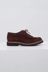 Zapato abotinado de gamuza color marrón, con cordones, detalle de flecos en el empeine y suela de goma negra con costura vista.