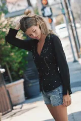 Camisa de cuello solapa con escote en pico y manga larga acabada en puño con abertura y lazo, confeccionada en tejido semitransparente de color negro con aplicación de tachas delanteras, cintura entallada con frunces laterales y cierre frontal con lazos.