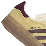 Championes adidas modelo Gazelle Bold, con diseño de plataforma de triple capa en color marrón claro. Presentan una capellada de gamuza en tono amarillo suave, con las icónicas tres tiras laterales y el refuerzo del talón en color bordó. Incluyen cordones blancos y lengüeta con textura grabada.