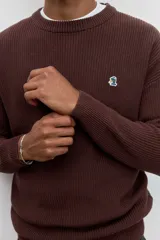 Sweater de tejido de punto acanalado en color marrón, con cuello redondo y ajuste holgado. Presenta un pequeño bordado distintivo en el pecho.