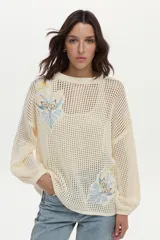 Sweater tejido en estructura calada, de color crudo, con mangas anchas, bordados decorativos en el frente y cinturón tejido incorporado.