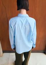 Camisa celeste de manga larga con diseño de rayas finas, cuello clásico, cierre frontal con botones y un bolsillo aplicado en el pecho.