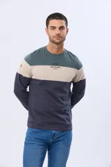 Remera de manga larga con cuello redondo y diseño de rayas horizontales en azul verdoso, beige y azul marino. Presenta una estampa frontal con texto y un parche decorativo en la manga.