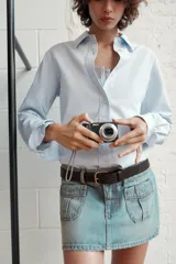 Falda mini de denim en tono azul claro, con tiro medio, trabillas para cinturón y bolsillos de plastrón en el frente. Presenta cierre frontal con cremallera y botones metálicos.