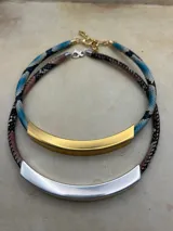 Collar de diseño minimalista con una pieza central curva de metal plateado mate y cordón trenzado con textura tipo piel de serpiente en tonos rosados y oscuros.