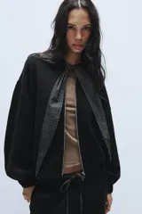 Chaqueta de punto negra con cuello redondo, cierre frontal con cremallera metálica y detalle de lazada en tejido a cuadros en contraste. Presenta mangas largas abullonadas con puños elásticos y bajo elástico.
