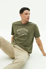 Remera verde militar de manga corta con cuello redondo y estampado frontal con el texto "Brookfield The Emporium of Postmodern Activity No 1001 Torino | 1971".
