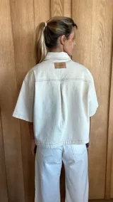 Sobrecamisa de denim soft en tono azul medio, con corte oversize, mangas cortas, cuello clásico y cierre frontal con botones metálicos. Presenta dos bolsillos frontales con solapa y un ruedo redondeado.