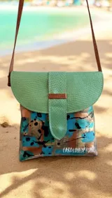 Cartera y bandolera de cuero Emi línea Ottenta, diseñada por Carolina de Cunto con arte pintado a mano. Presenta una solapa semicircular de cuero verde con textura símil serpiente y broche imán, cuerpo principal con estampado artístico multicolor en tonos celestes, marrones y negros, y correa larga marrón fina.