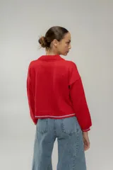 Cardigan de punto color rojo, con cuello redondo, cierre frontal de botones y mangas largas. Presenta un detalle bordado de una concha marina en el pecho y terminaciones en contraste de color celeste en el ruedo, puños y bolsillo frontal.