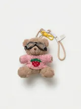 Llavero de peluche en forma de osito, vestido con un suéter tejido rosado con detalle de fresa bordada y gafas de aviador. Cuenta con correa de muñeca beige y ganchos metálicos y plásticos.